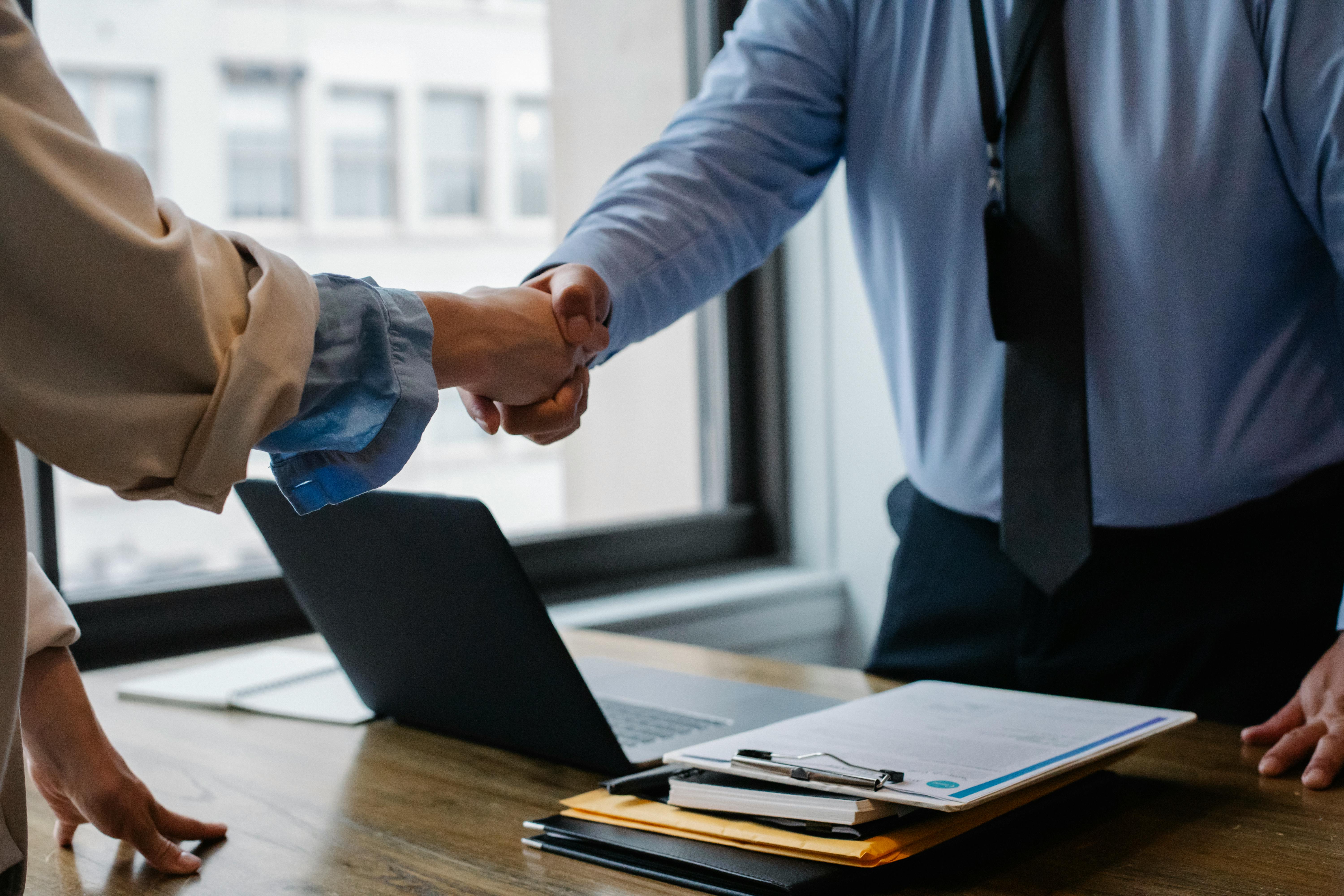 People shaking hands over a desk that has a laptop on it